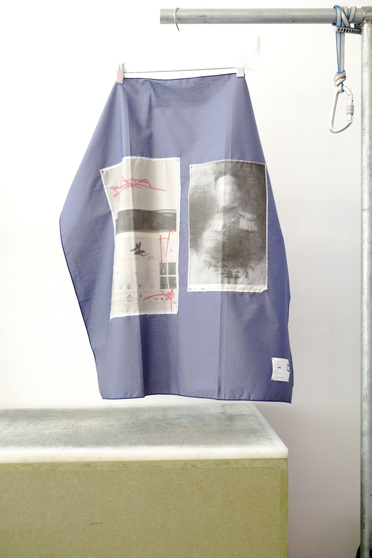 A blue foulard with two print panels hanging from a metal hanger on a clothing rack, positioned above a concrete block against a white wall.