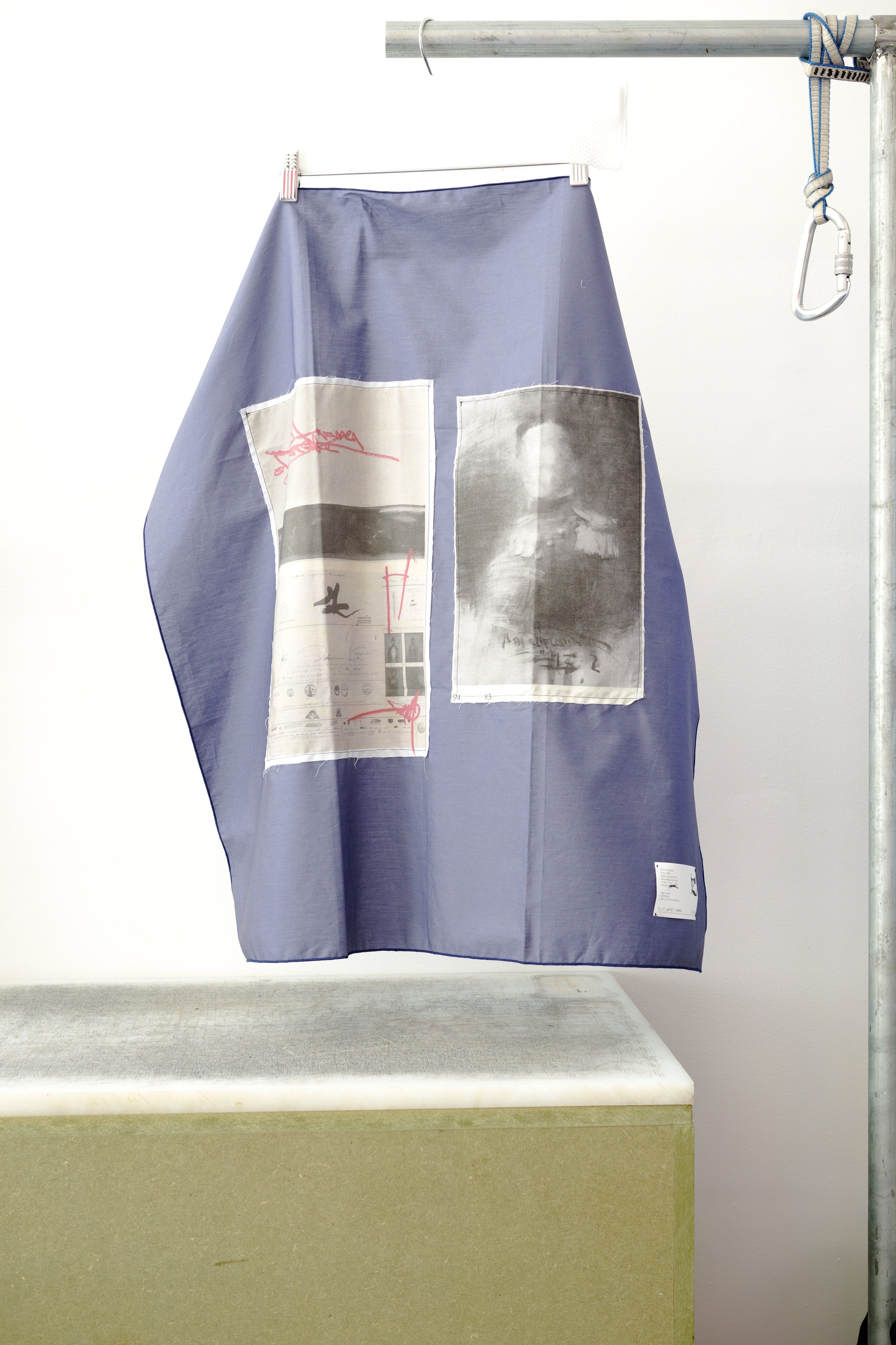 A blue foulard with two print panels hanging from a metal hanger on a clothing rack, positioned above a concrete block against a white wall.