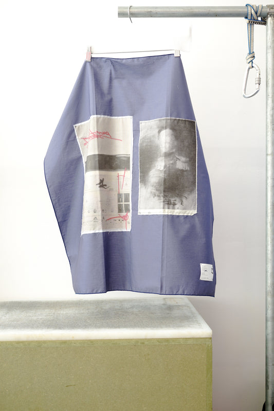 A blue foulard with two print panels hanging from a metal hanger on a clothing rack, positioned above a concrete block against a white wall.