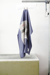 A blue foulard with two print panels hanging from a metal hanger on a clothing rack, positioned above a concrete block against a white wall.