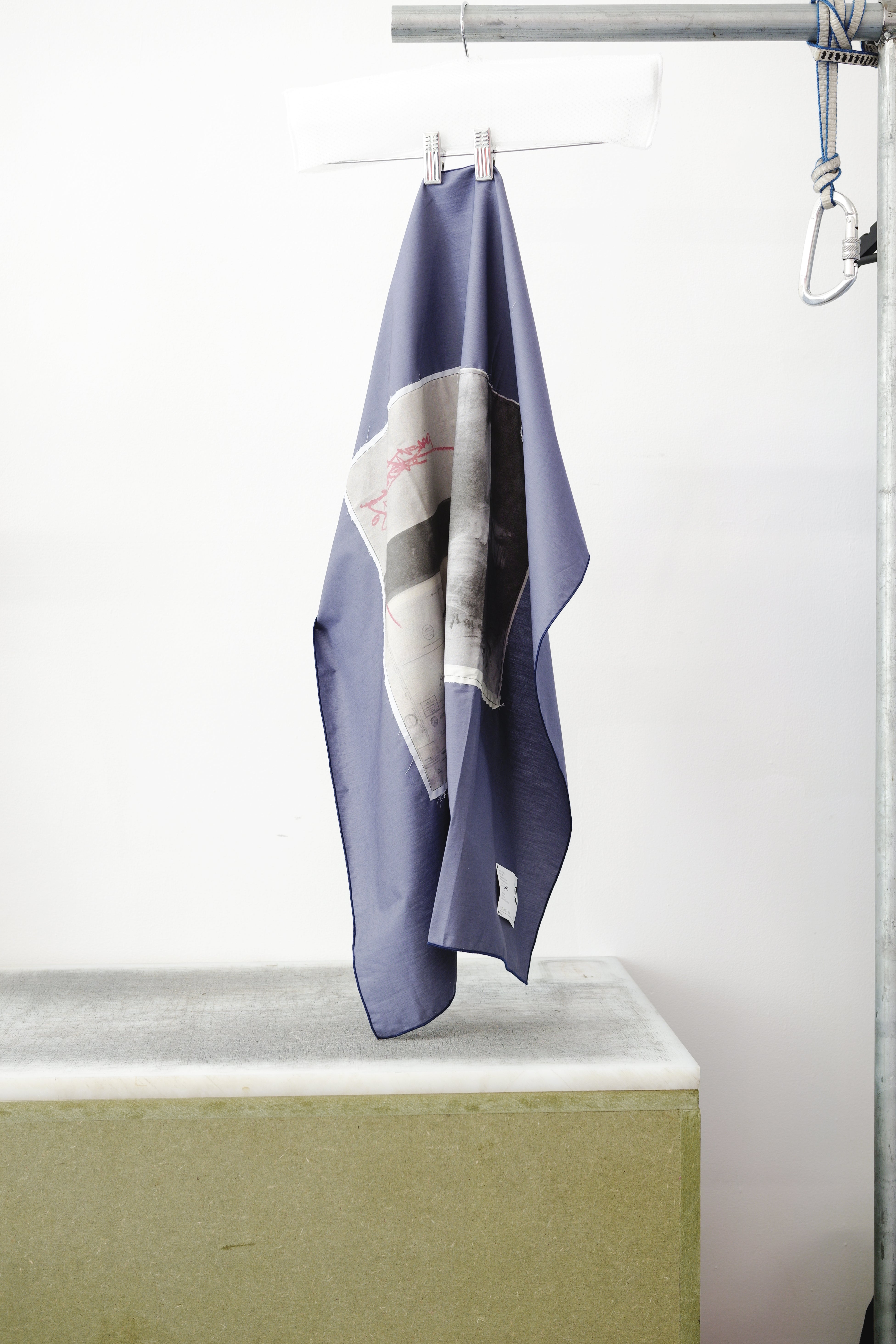 A blue foulard with two print panels hanging from a metal hanger on a clothing rack, positioned above a concrete block against a white wall.