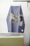 A blue foulard with burgandy grid and two print panels hanging from a metal hanger on a clothing rack, positioned above a concrete block against a white wall.