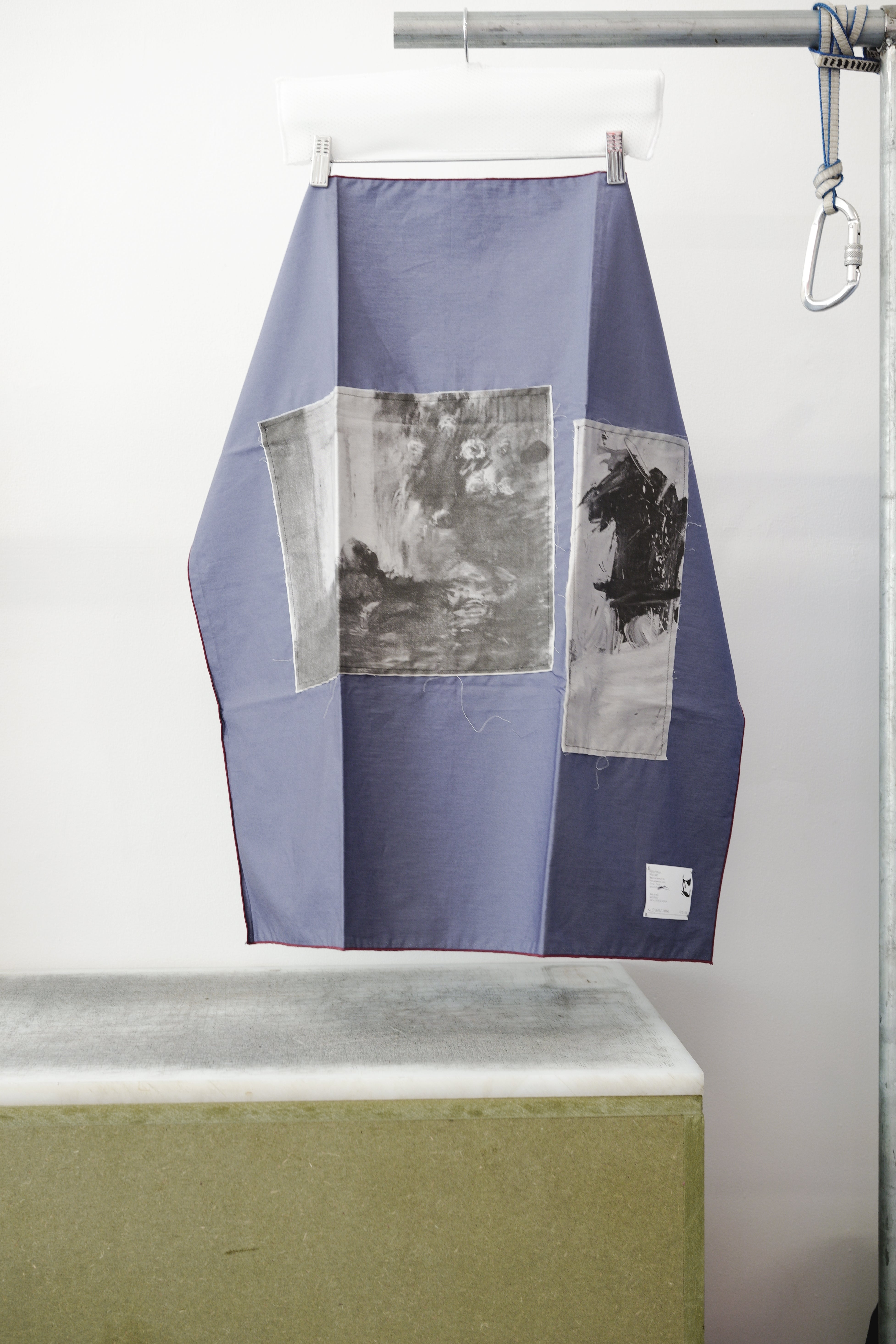 A blue foulard with burgandy grid and two print panels hanging from a metal hanger on a clothing rack, positioned above a concrete block against a white wall.