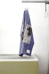 A blue foulard with burgandy grid and two print panels hanging from a metal hanger on a clothing rack, positioned above a concrete block against a white wall.