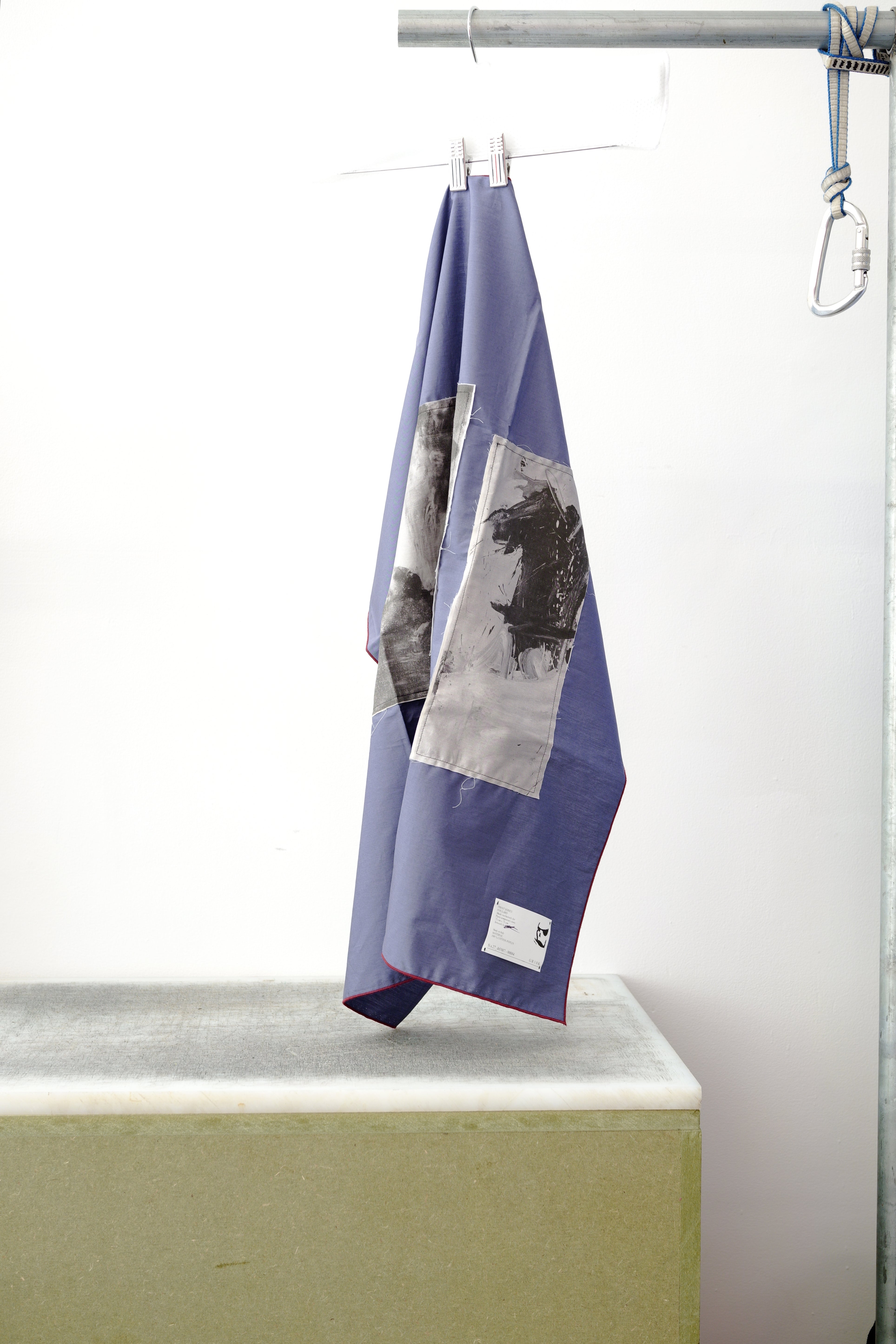 A blue foulard with burgandy grid and two print panels hanging from a metal hanger on a clothing rack, positioned above a concrete block against a white wall.