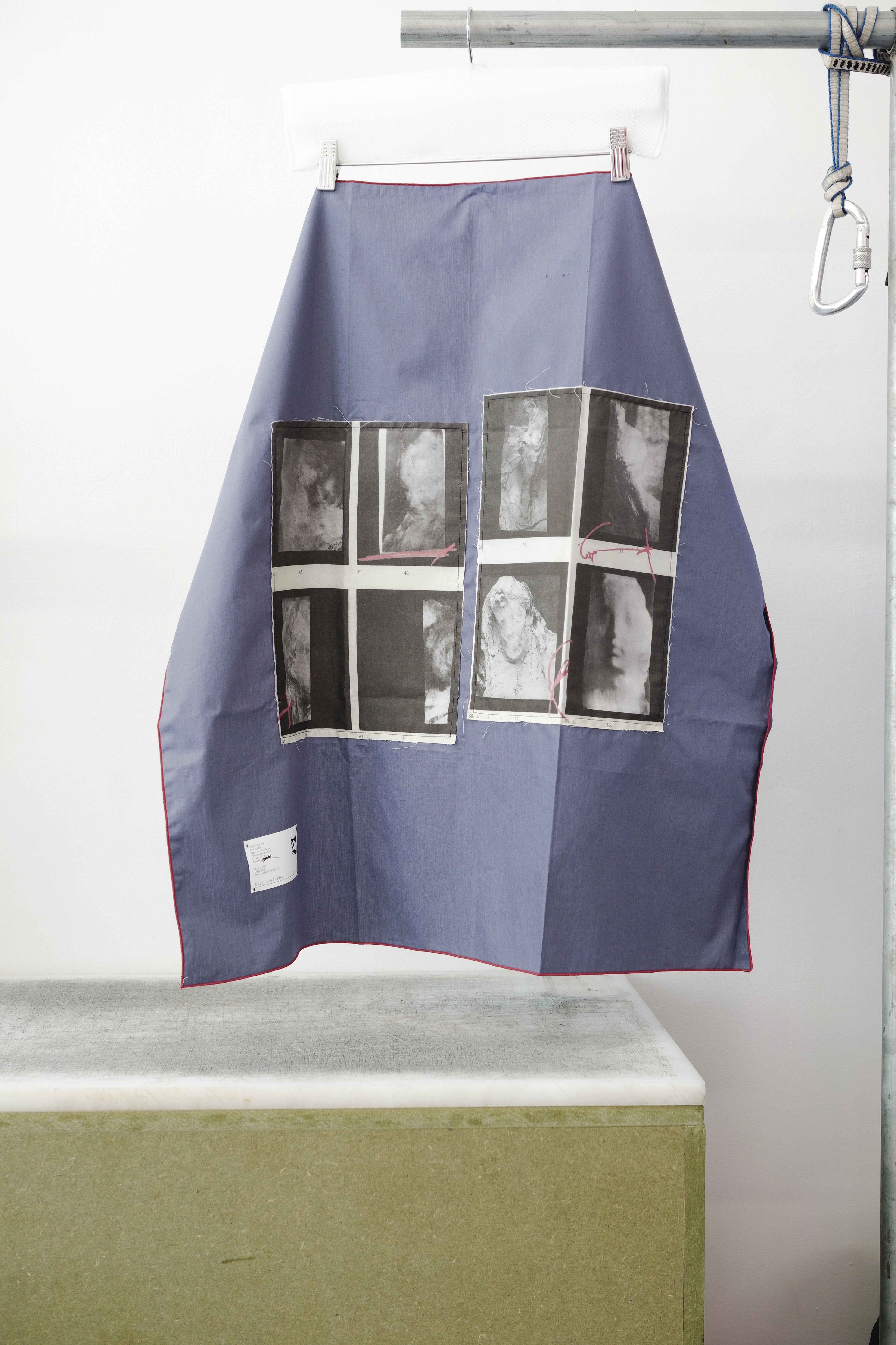 A blue foulard with red grid and two print panels hanging from a metal hanger on a clothing rack, positioned above a concrete block against a white wall.