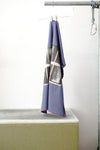 A blue foulard with burgandy grid and two print panels hanging from a metal hanger on a clothing rack, positioned above a concrete block against a white wall.