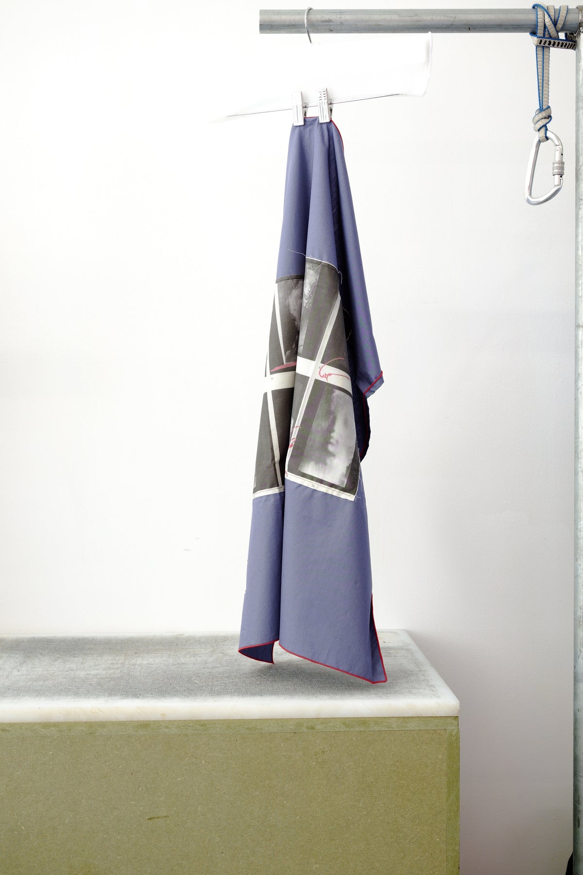 A blue foulard with burgandy grid and two print panels hanging from a metal hanger on a clothing rack, positioned above a concrete block against a white wall.