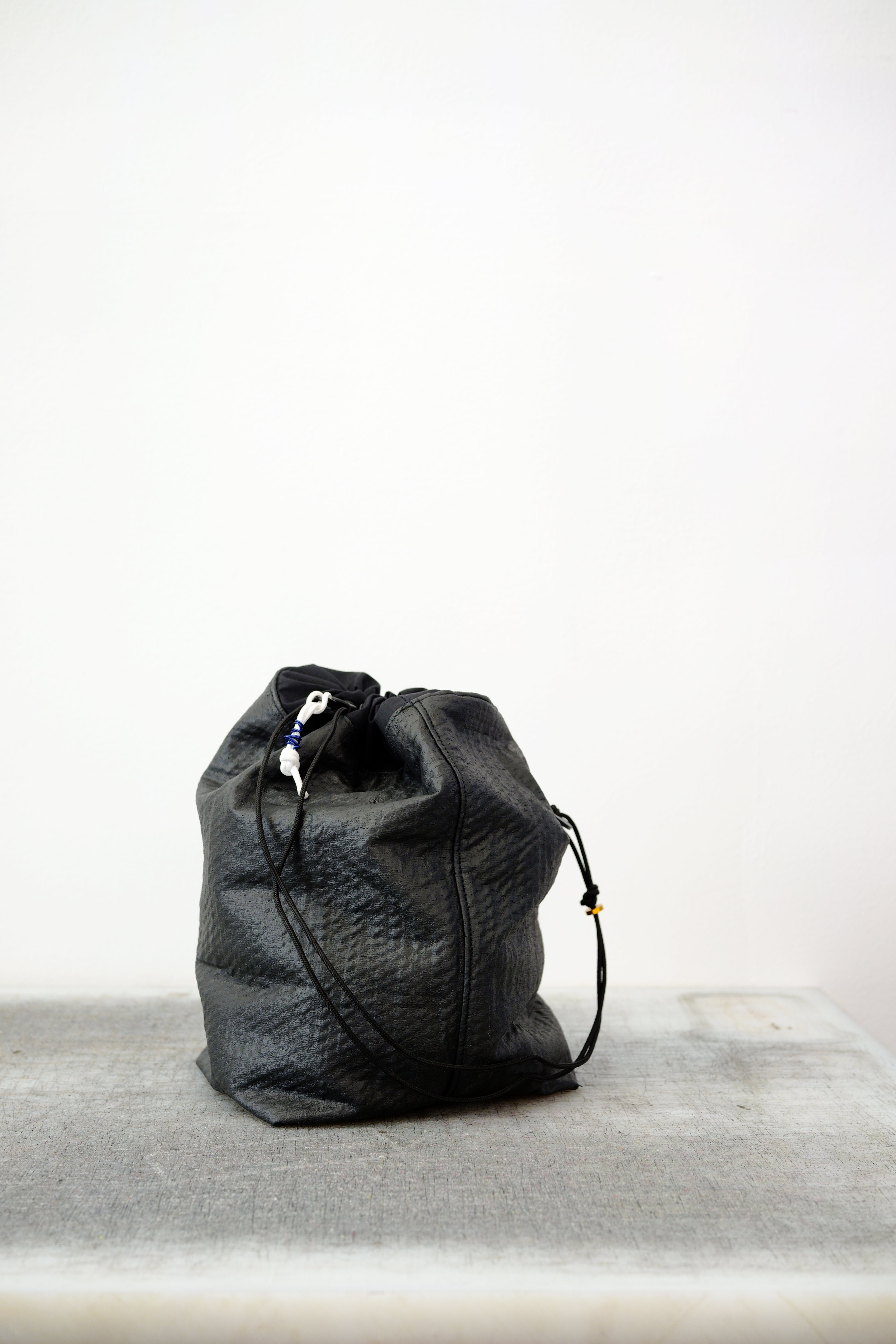 Black drawstring bag with white drawstrings and GR10K logo patch, against a plain white wall.