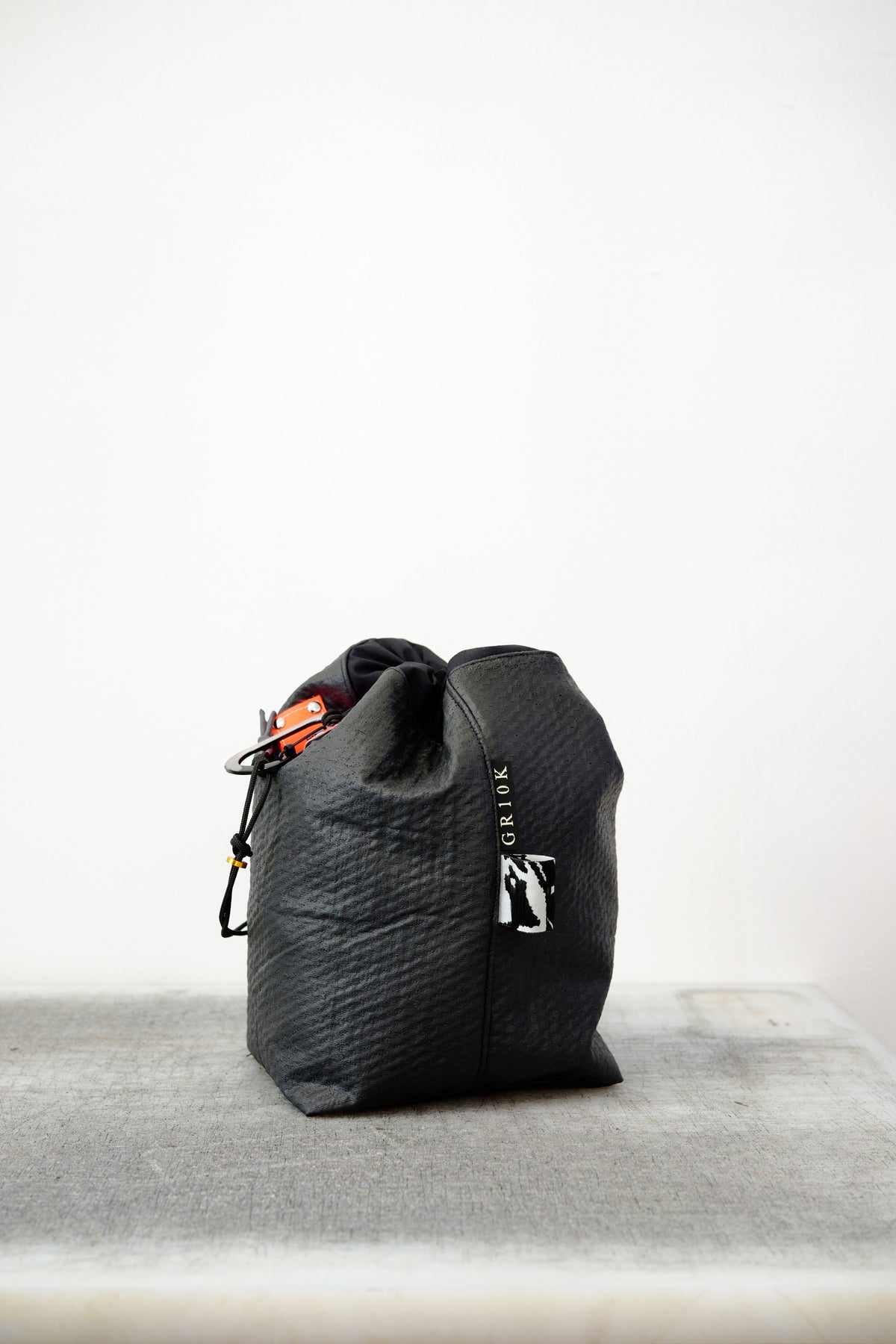 Black drawstring bag with white drawstrings and GR10K logo patch, against a plain white wall.