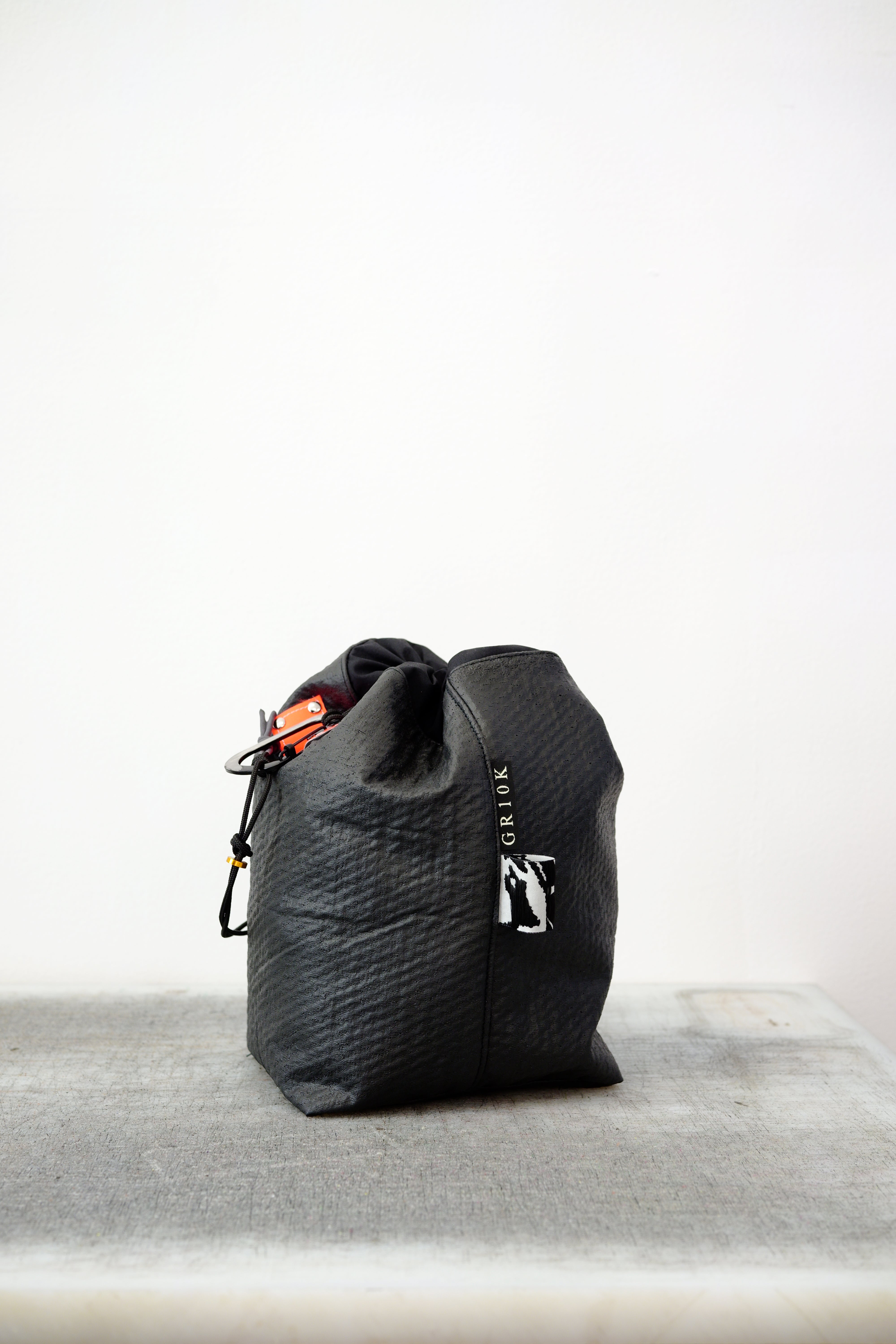 Black drawstring bag with white drawstrings and GR10K logo patch, against a plain white wall.
