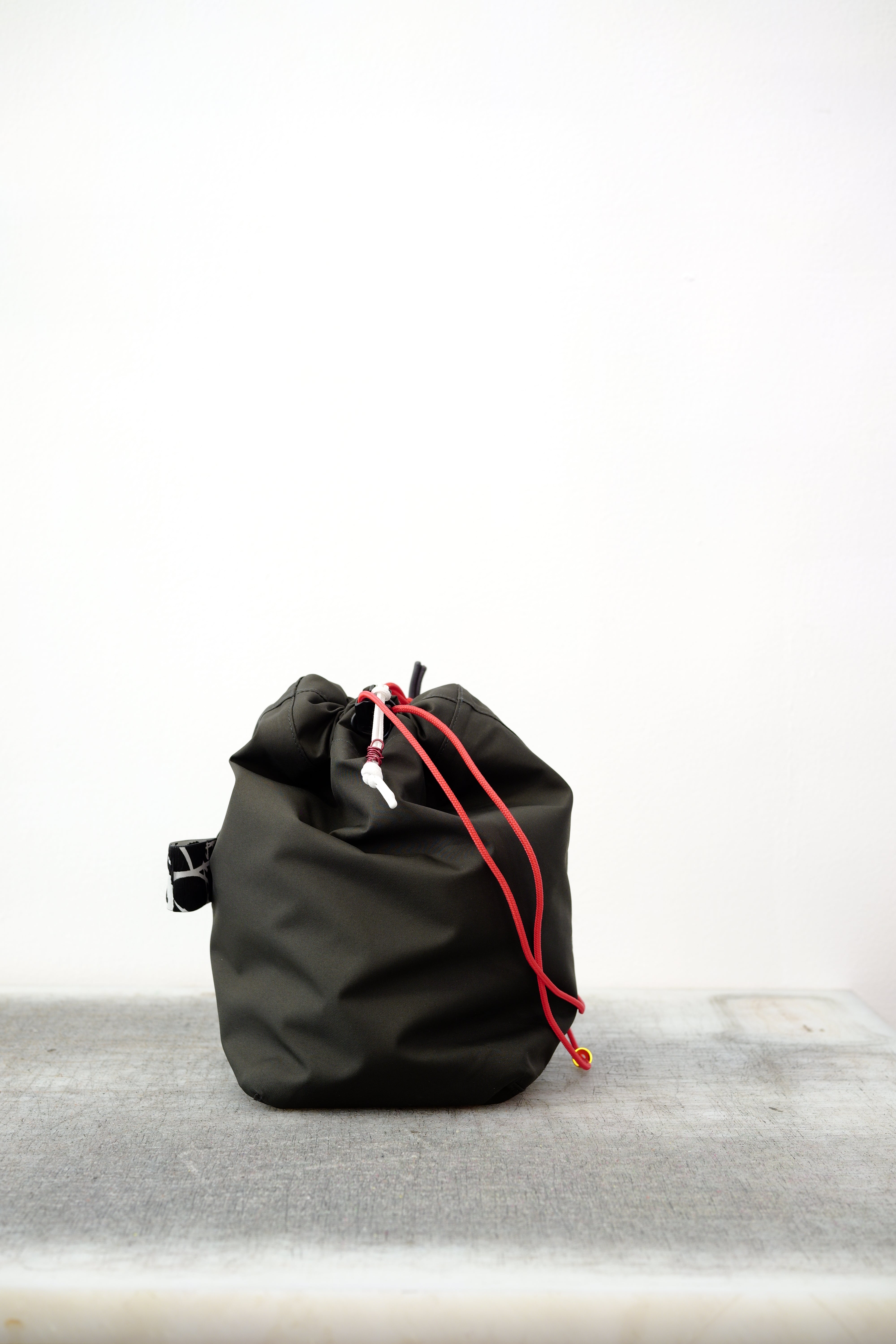 Black drawstring bag with red drawstrings and GR10K logo patch, against a plain white wall.