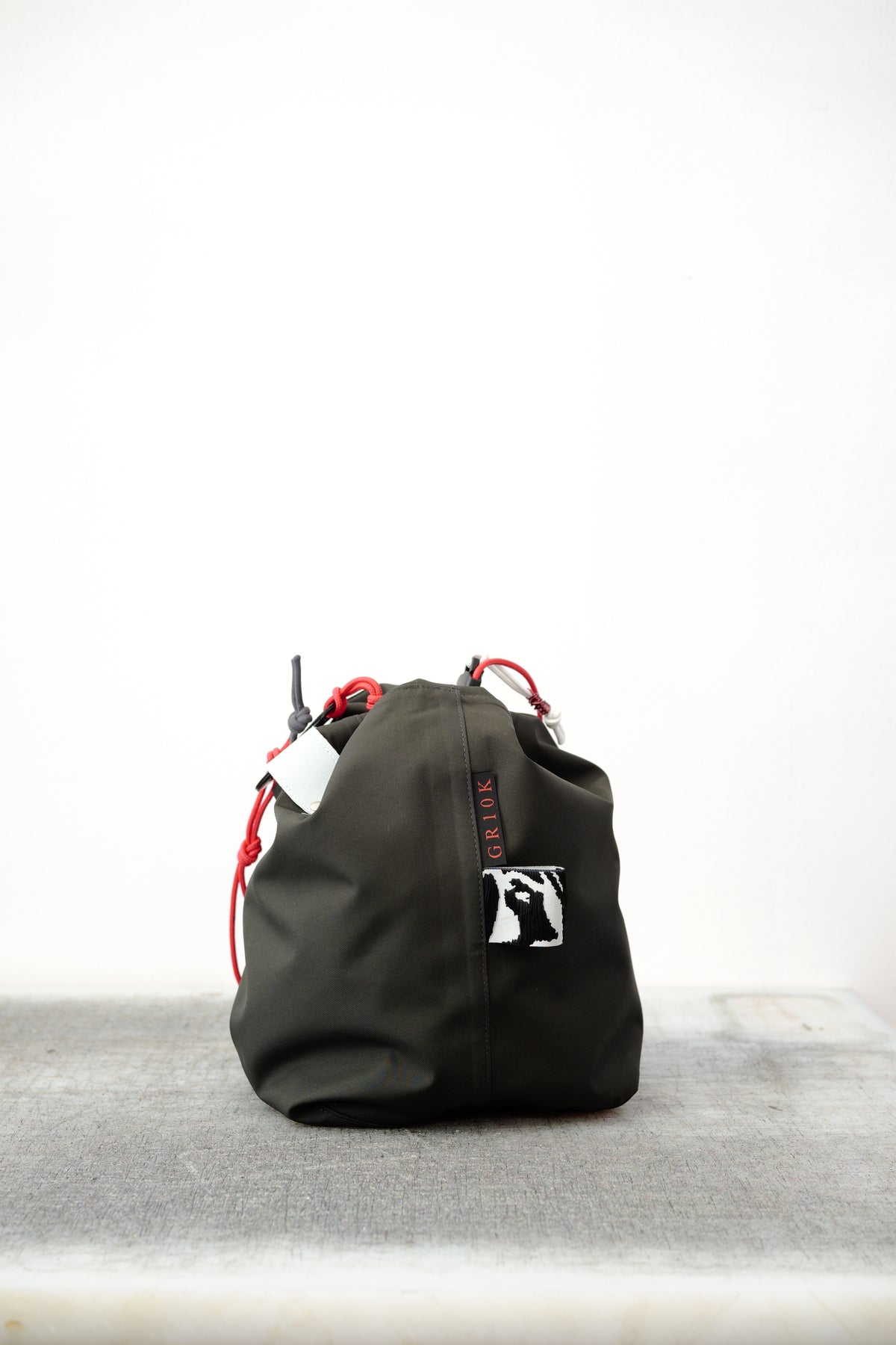 Black drawstring bag with red drawstrings and GR10K logo patch, against a plain white wall.