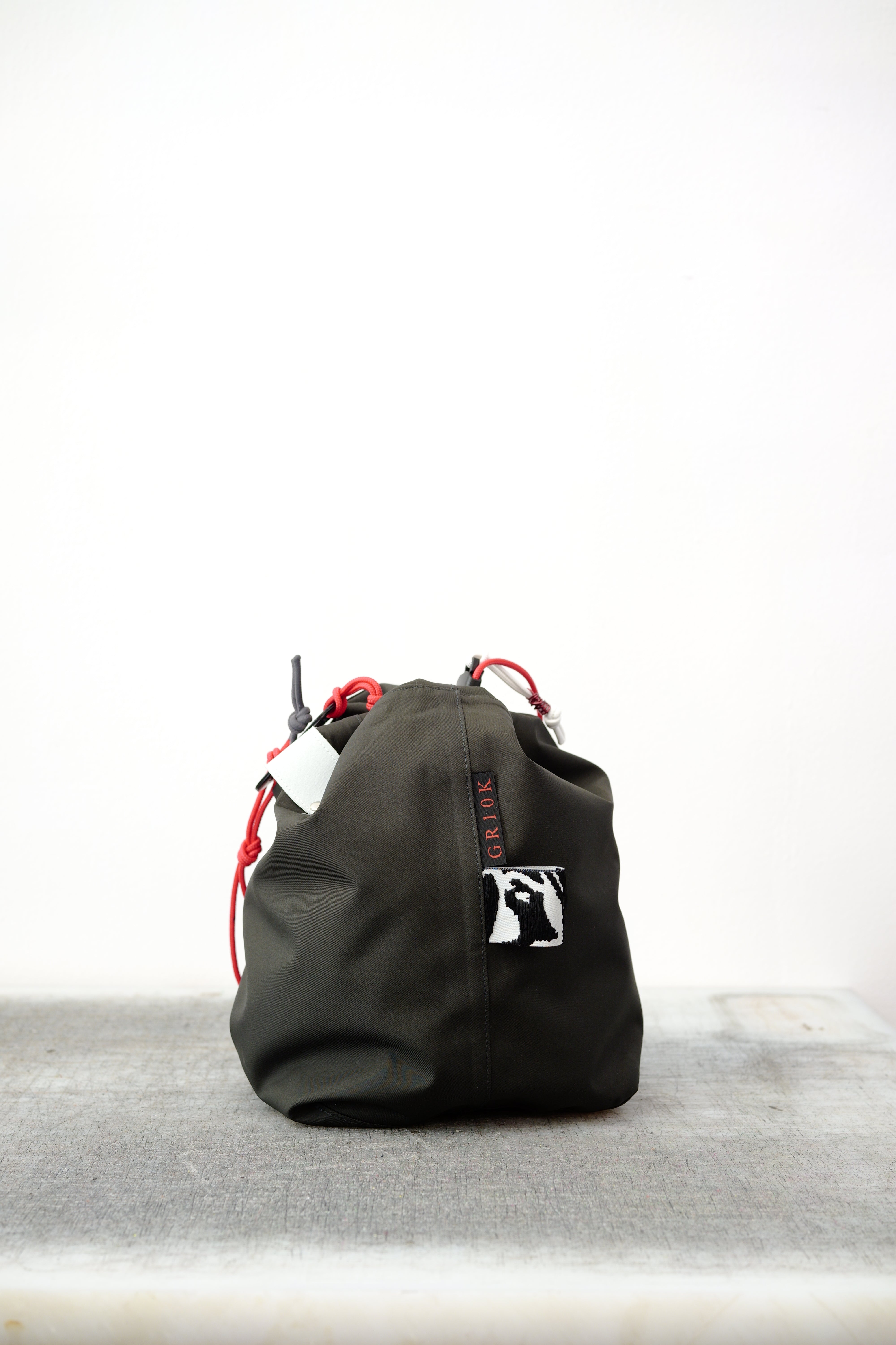 Black drawstring bag with red drawstrings and GR10K logo patch, against a plain white wall.