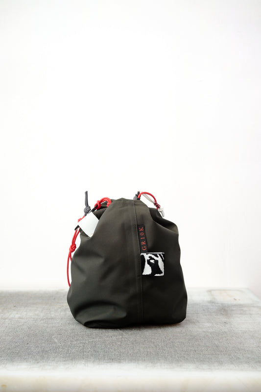 Black drawstring bag with red drawstrings and GR10K logo patch, against a plain white wall.