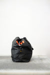 Black drawstring bag with white drawstrings and GR10K logo patch, against a plain white wall.