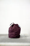 Burgundy drawstring bag with white drawstrings and GR10K logo patch, against a plain white wall.