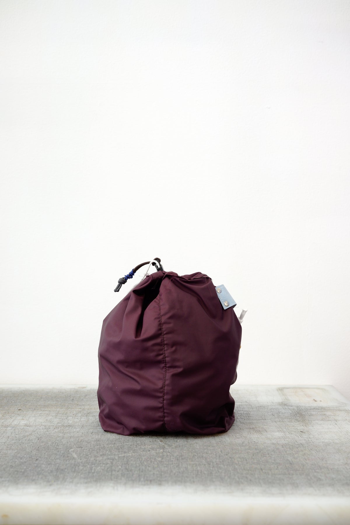 Burgundy drawstring bag with white drawstrings and GR10K logo patch, against a plain white wall.