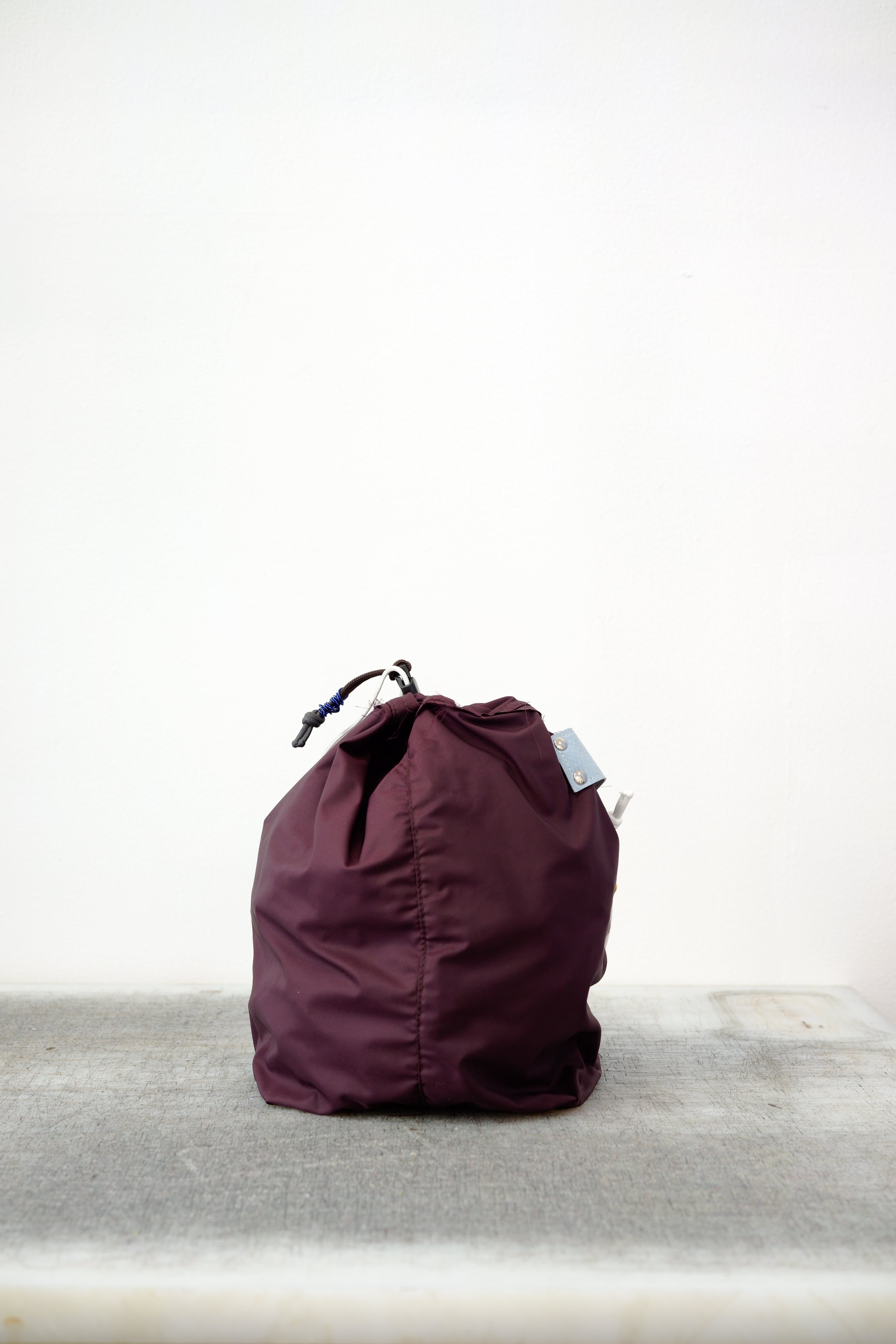 Burgundy drawstring bag with white drawstrings and GR10K logo patch, against a plain white wall.