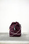 Burgundy drawstring bag with white drawstrings and GR10K logo patch, against a plain white wall.