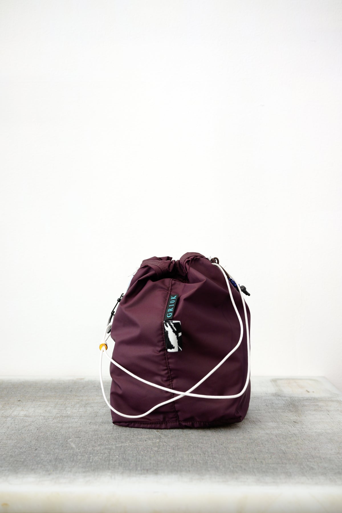 Burgundy drawstring bag with white drawstrings and GR10K logo patch, against a plain white wall.