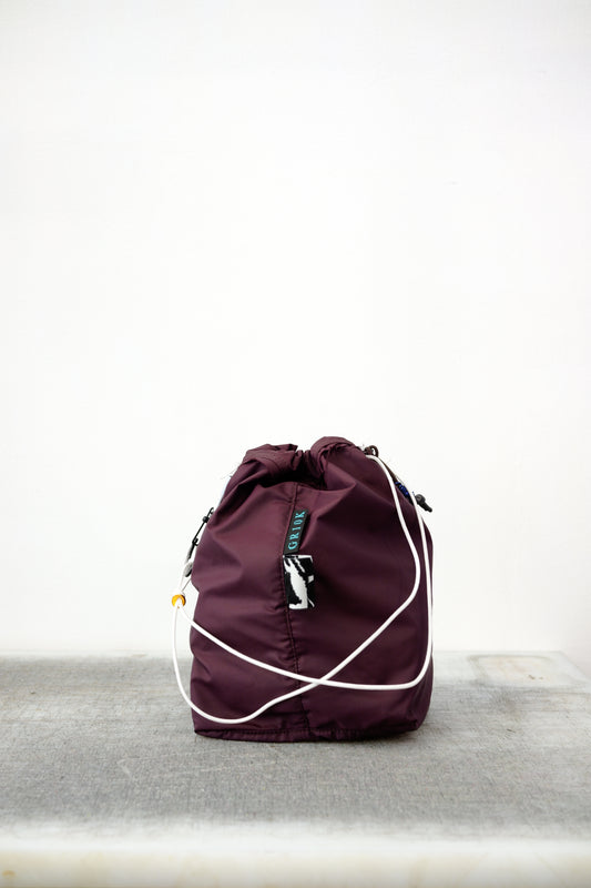 Burgundy drawstring bag with white drawstrings and GR10K logo patch, against a plain white wall.