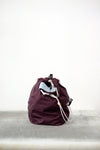 Burgundy drawstring bag with white drawstrings and GR10K logo patch, against a plain white wall.