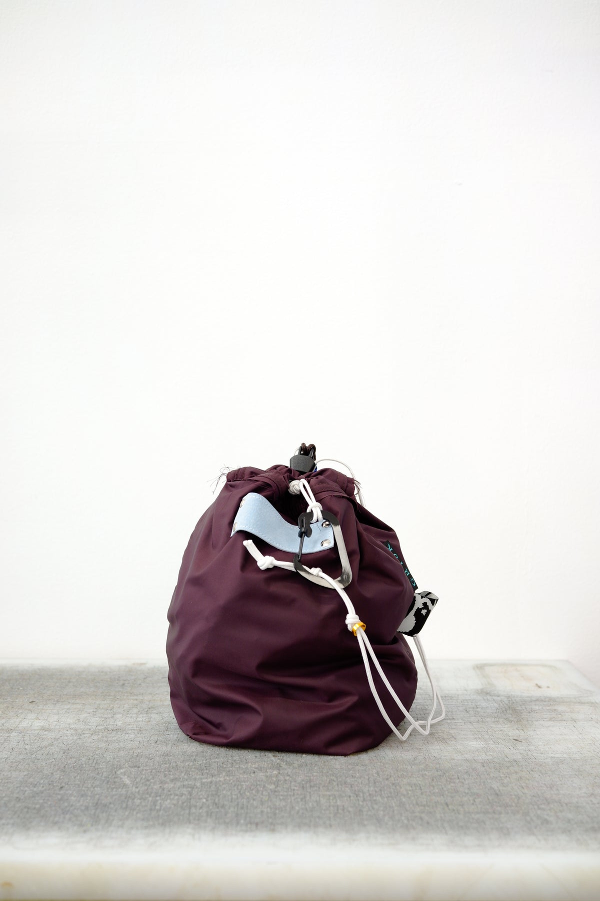 Burgundy drawstring bag with white drawstrings and GR10K logo patch, against a plain white wall.
