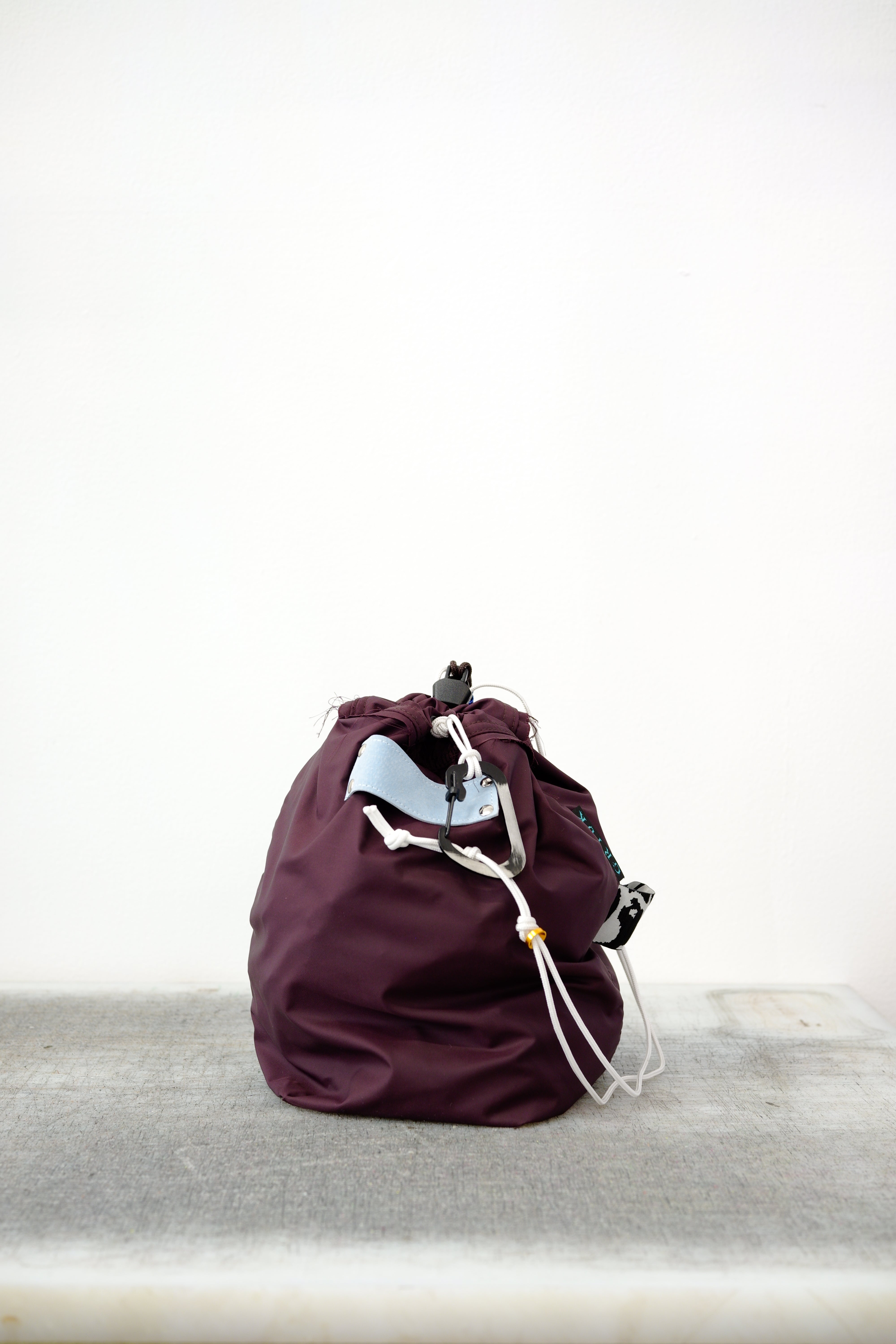 Burgundy drawstring bag with white drawstrings and GR10K logo patch, against a plain white wall.