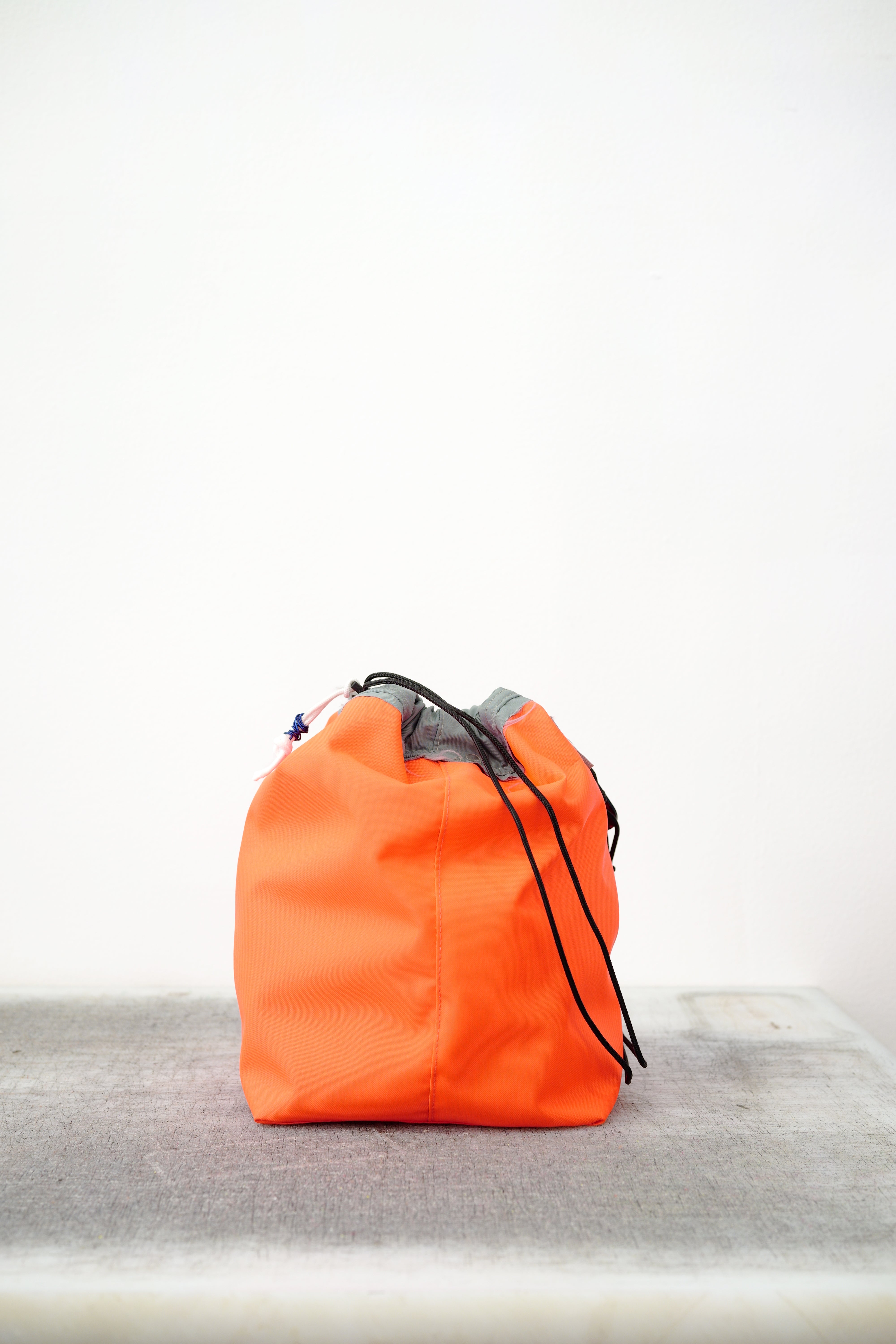 Orange drawstring bag with white drawstrings and GR10K logo patch, against a plain white wall.