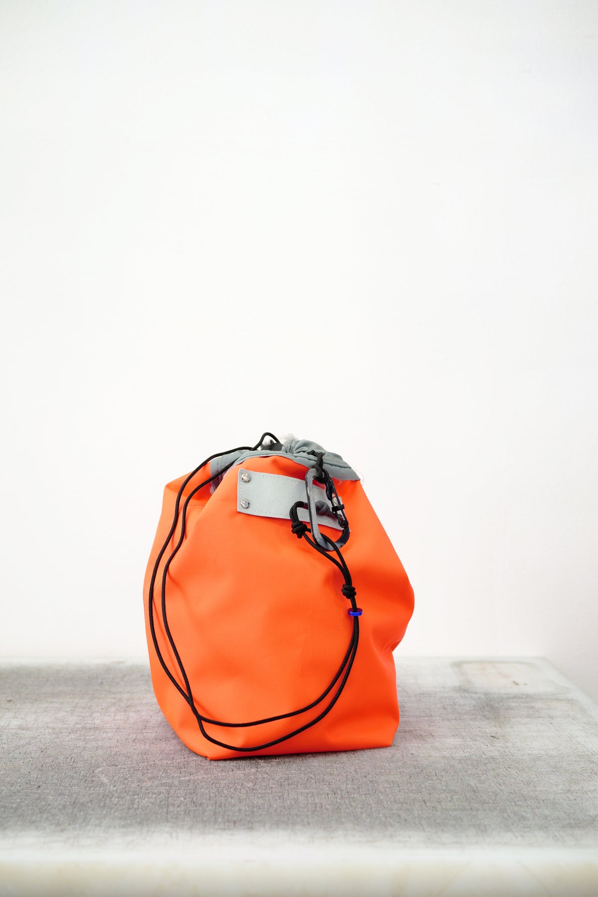 Orange drawstring bag with white drawstrings and GR10K logo patch, against a plain white wall.
