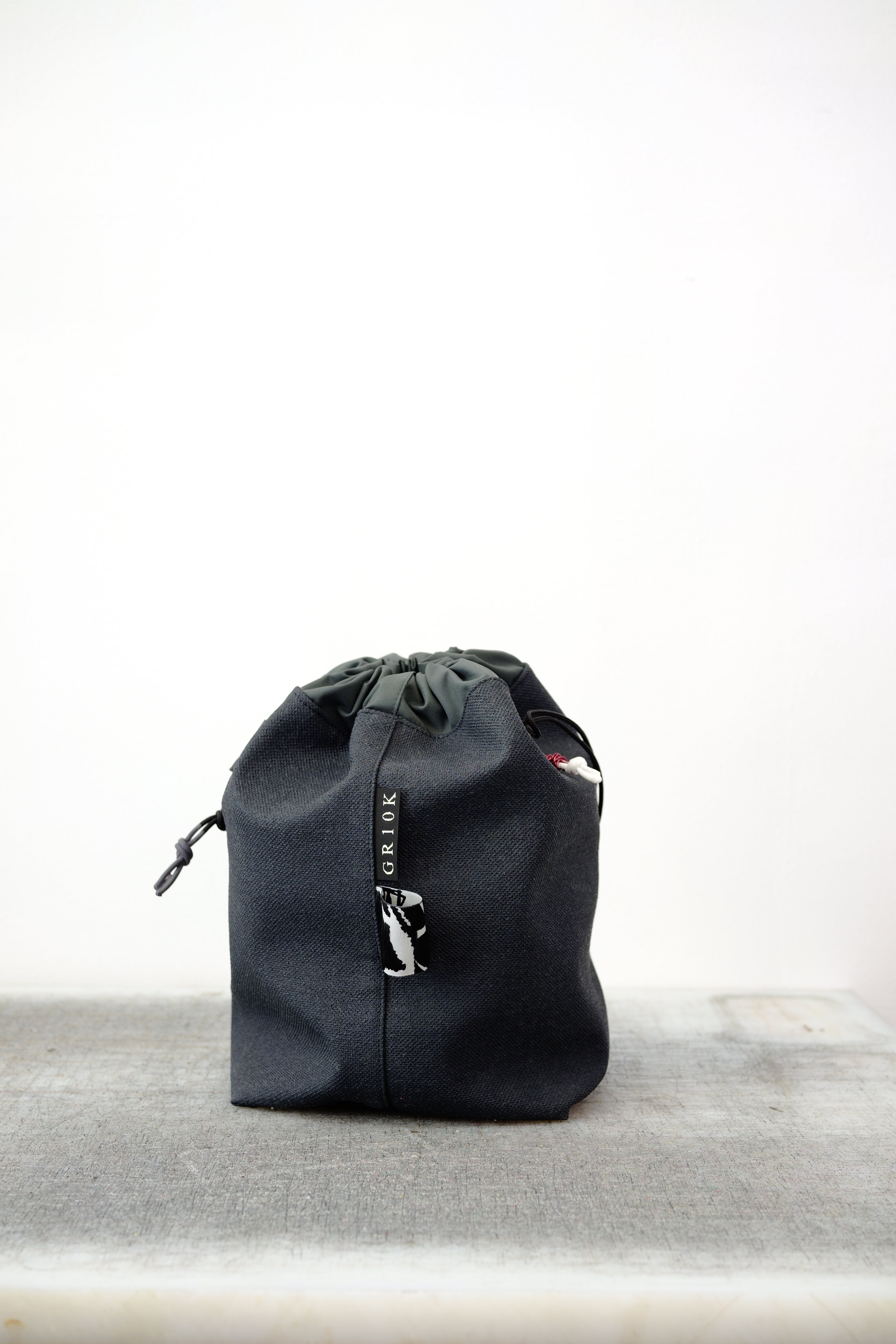 Grey drawstring bag with black drawstrings and GR10K logo patch, against a plain white wall.