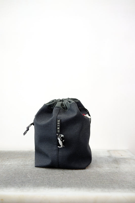 Grey drawstring bag with black drawstrings and GR10K logo patch, against a plain white wall.