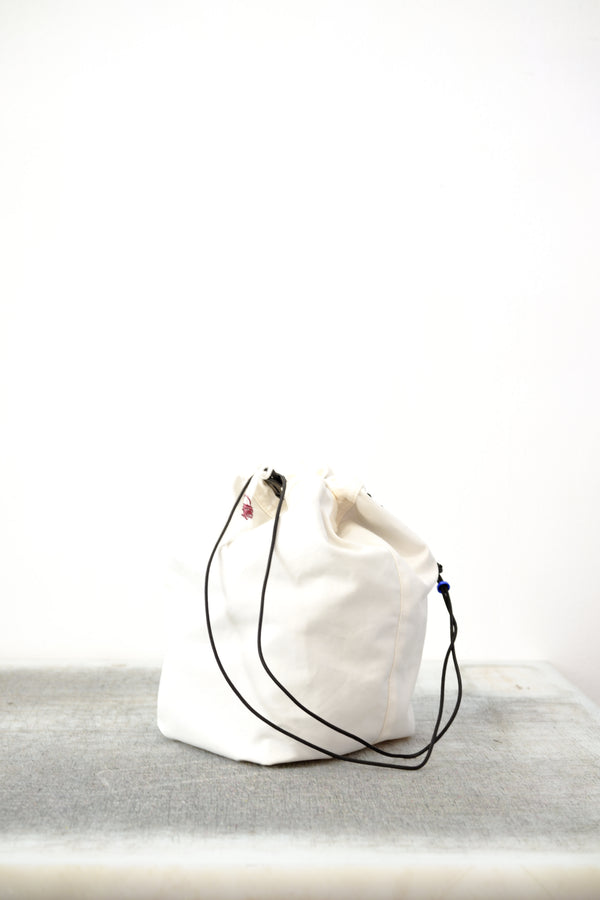 White drawstring bag with white drawstrings and GR10K logo patch, against a plain white wall.