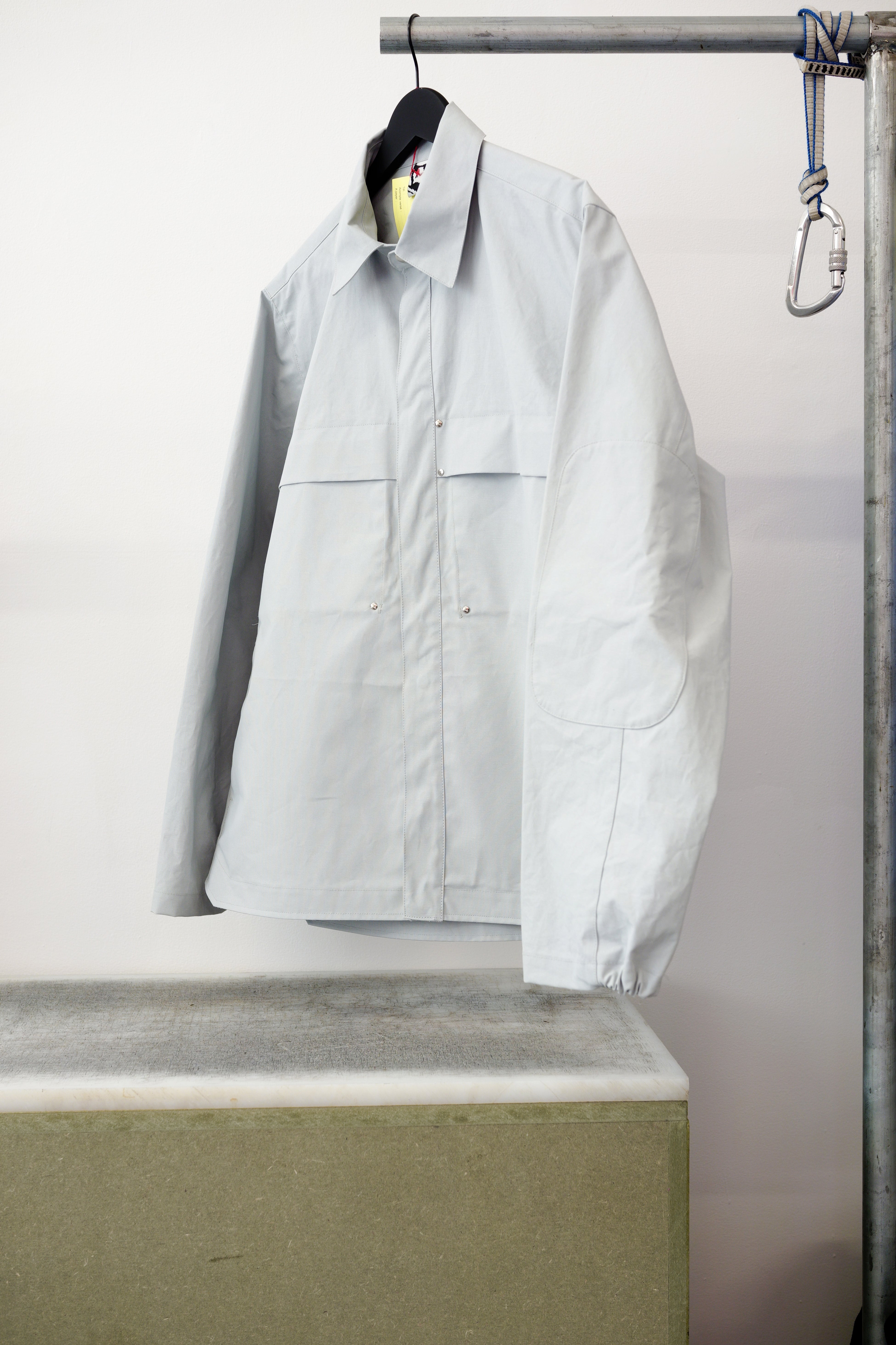 Gray utility overshirt with a pointed collar, button front, and two large chest flap pockets, hanging on a black hanger against white background
