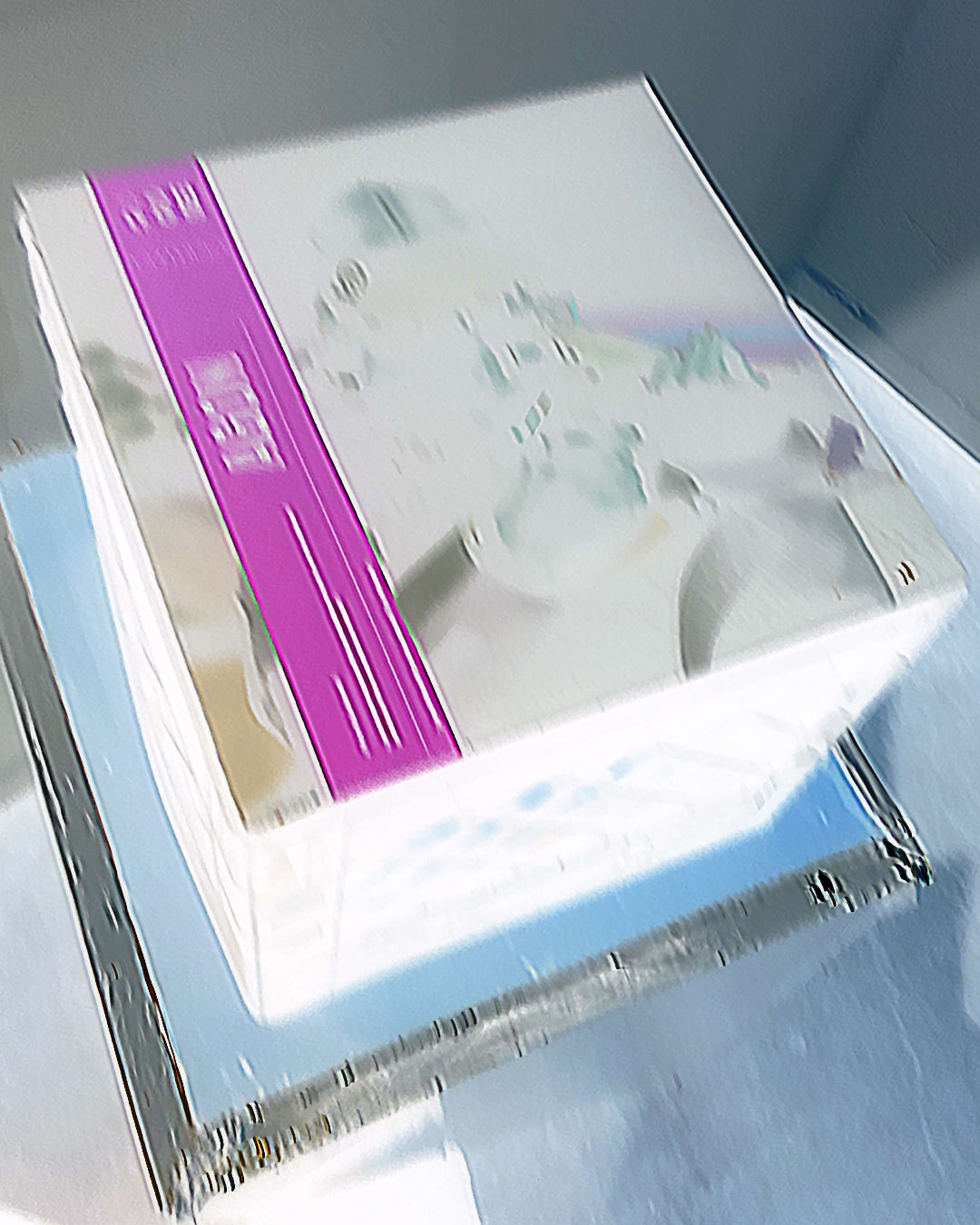 A boxed product photographed at an angle on a reflective silver tray. The box is predominantly white with a wide vertical purple band and soft pastel abstract artwork on the top.