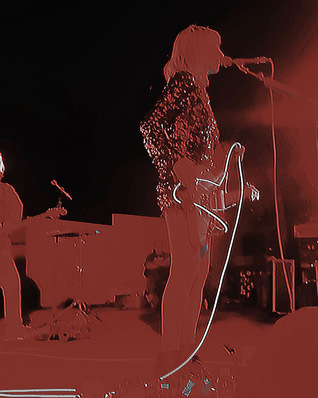 A high-contrast red-toned photograph of a live music performer on stage, holding a microphone and guitar, with other band equipment blurred in the background.