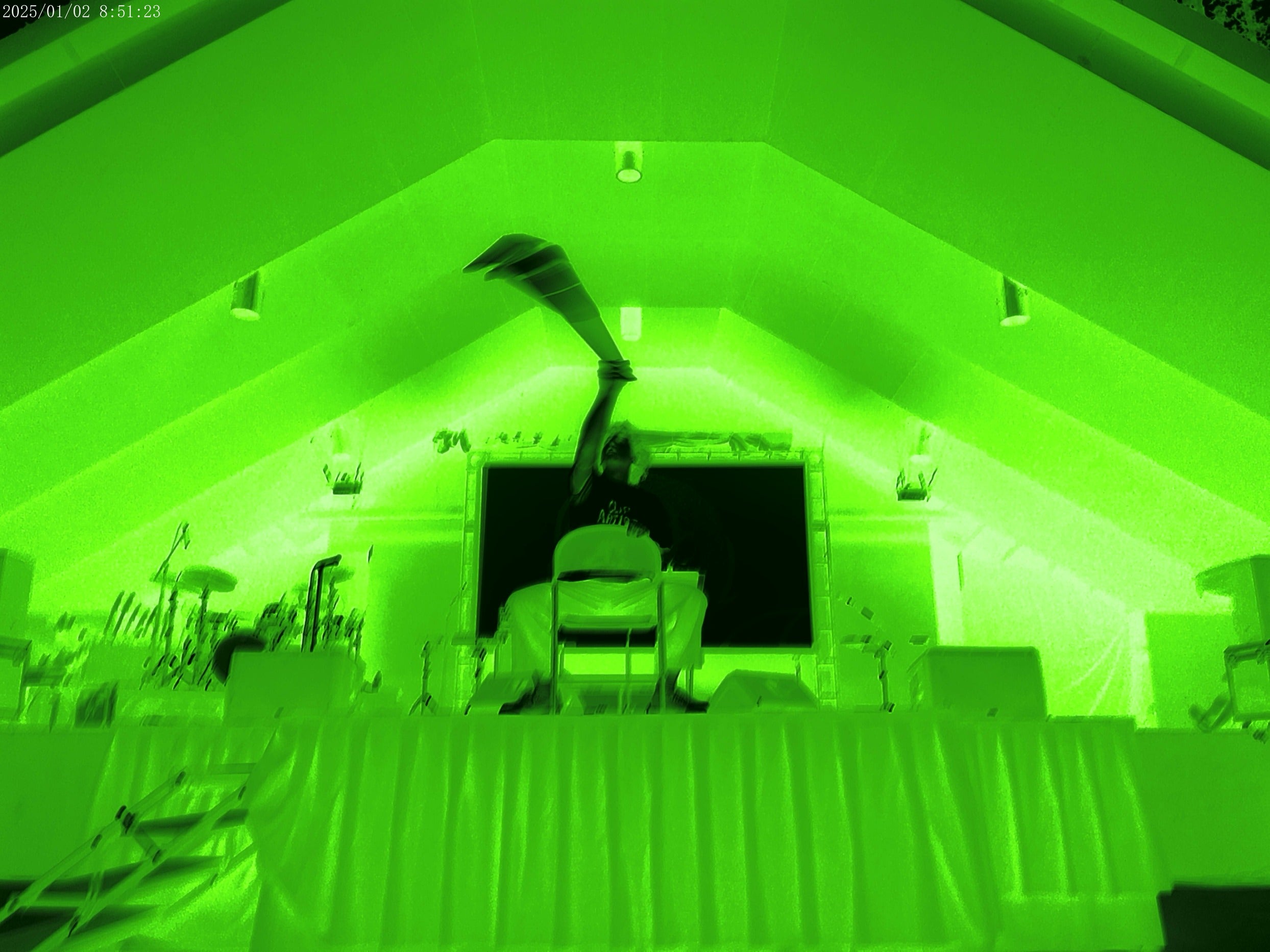 A green-tinted photo showing a performance on a stage with a long object extended upward, inside a tent-like structure with draped table in front.