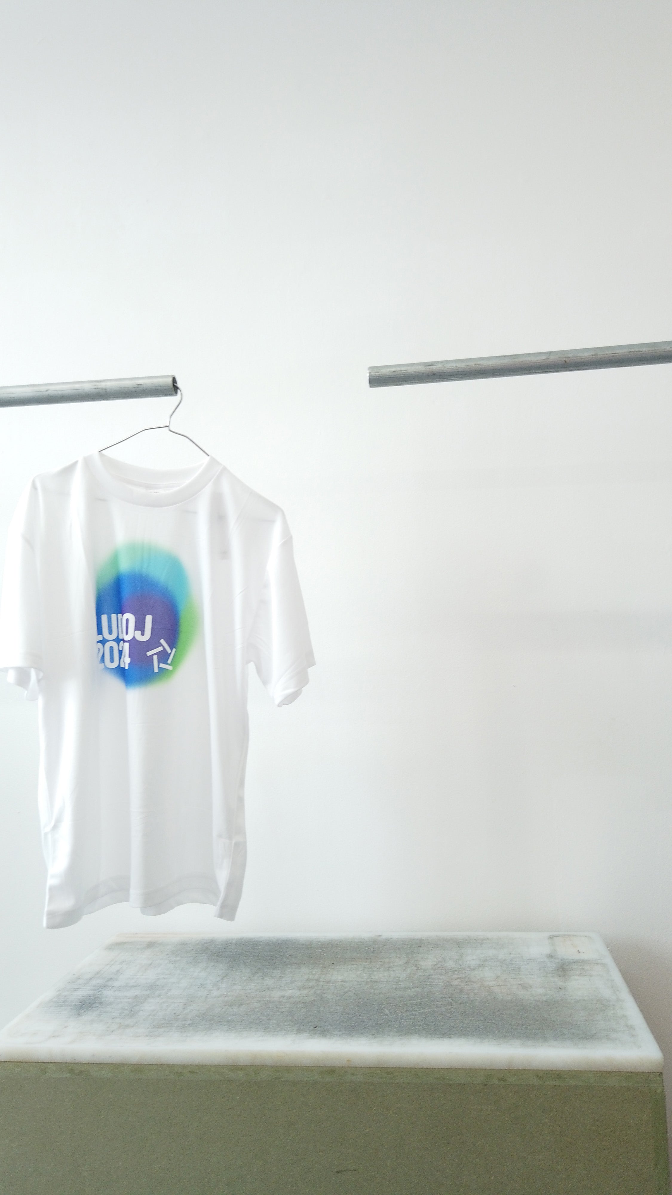 White short-sleeve T-shirt hanging on a metal rack against a plain white wall, featuring a circular blue and green graphic.