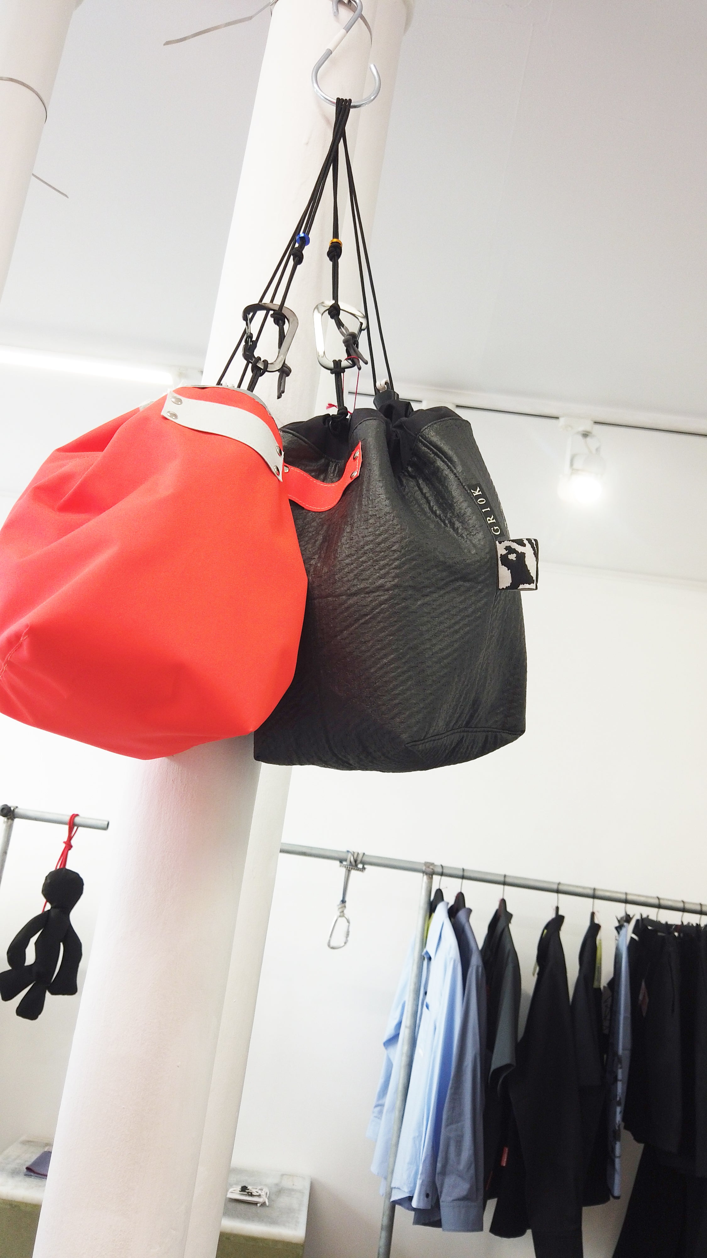 Two drawstring bags hanging from a ceiling hook in a retail store: a bright red bag on the left and a black textured bag on the right, with clothing racks visible in the background.