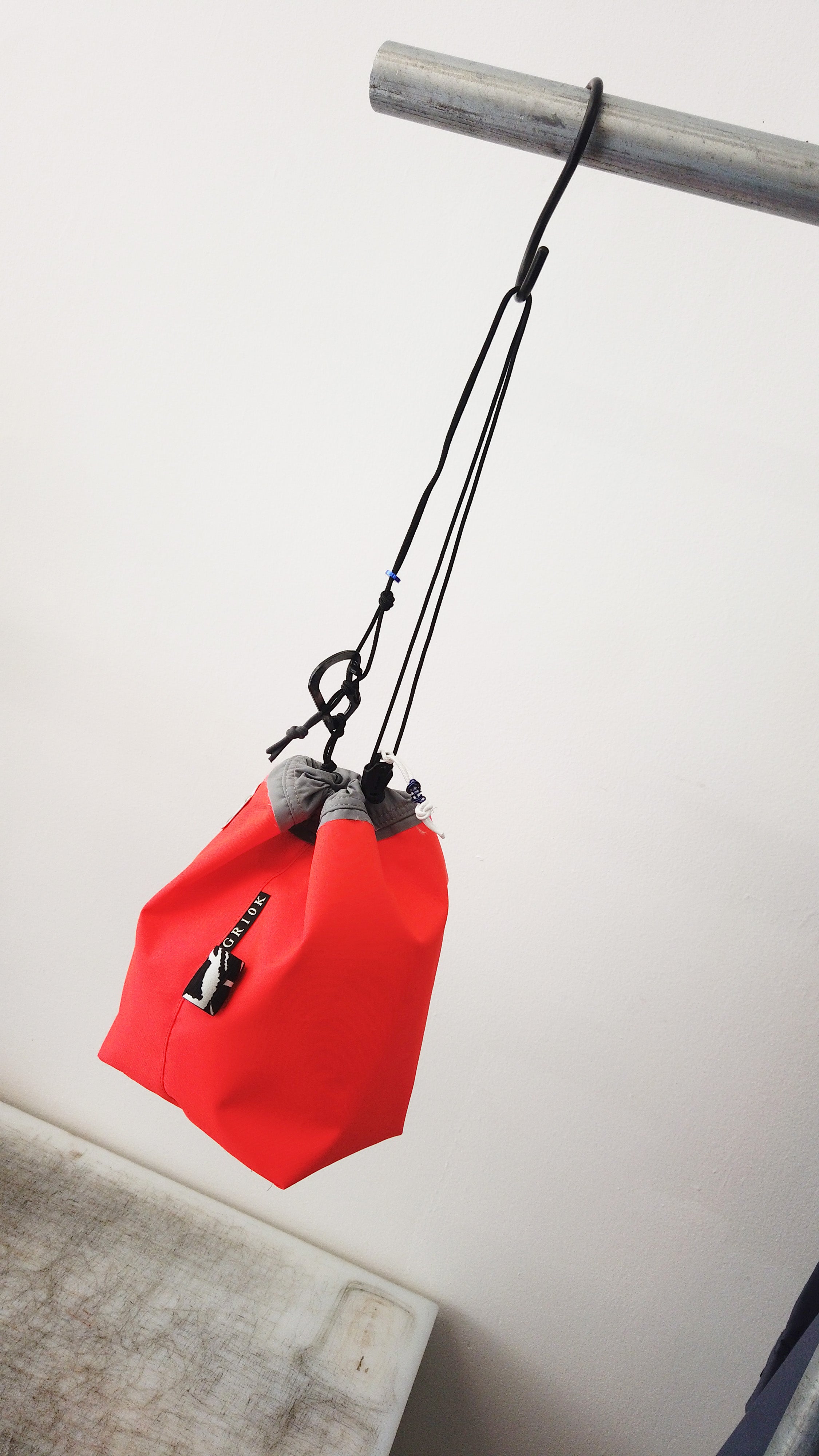 A bright red stuff sack with a black zipper and gray drawstring cords, hanging from a metal bar against a plain white wall.
